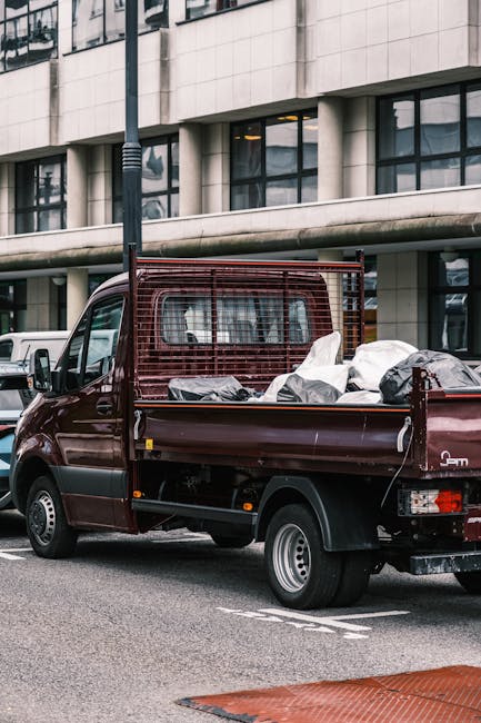 A dark red, medium-sized flatbed truck parked on a city street in front of a modern office building with large windows and concrete facade. The truck's flatbed is loaded with various bulky waste items, including large white and black plastic bags, cardboard boxes, and wrapped furniture pieces, all arranged towards the rear of the vehicle. The flatbed has a red metal mesh side panel for safety, and the items appear ready for disposal or recycling, indicating a waste clearance or home relocation process. In the background, other vehicles are visible parked along the street, and a street lamp is positioned near the truck. The scene is under natural daylight, with the environment suggesting an urban setting suitable for a professional removals service like Man with Van Hampton, specializing in house removals and waste disposal during moves.