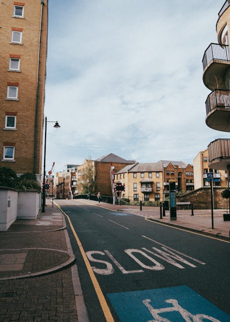 A street scene showing the exterior of several multi-storey residential buildings with brick and concrete facades, balconies, and windows. The road curves slightly and is marked with a large 'SLOW' sign painted in white on the asphalt, with a blue marked parking or loading zone nearby. There is a tall street lamp on the left side of the image, and a few pedestrians are visible walking along the pavement. In the foreground, a man with a moving box wrapped in plastic and cardboard stands on the pavement near the entrance of one building. Man with Van Hampton, a professional removal company, is preparing for a home relocation in Hampton's walk-up buildings, with furniture, boxes, and packing materials visible as part of the loading process inside the property and on the pavement adjacent to a transport van, which is partially out of the frame on the right side. The scene is captured during daylight with overcast skies, utilizing natural lighting to showcase elements of furniture transport, packing, and loading steps involved in house removals.