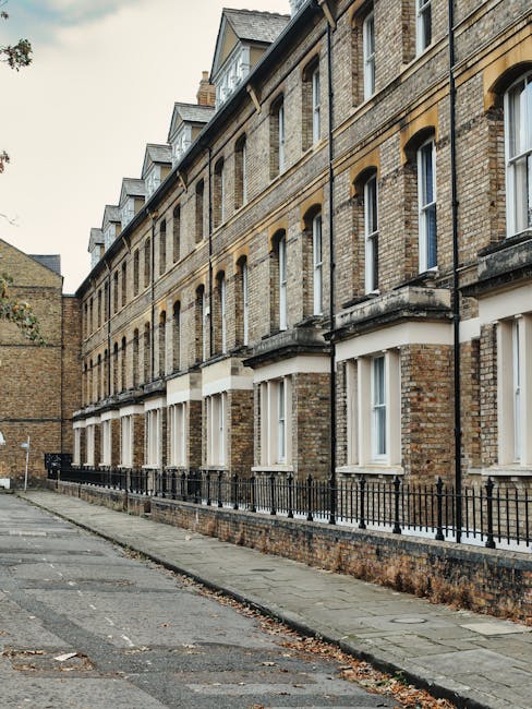 The image shows a row of four-storey brick residential buildings with large ground-floor windows framed in white. The buildings feature small, decorative balconies with black metal railings and a sloped slate roof with dormer windows on the upper floor. The street in front is paved with concrete, with a narrow sidewalk and a low brick wall topped by a black metal fence running along the front of the buildings. In the foreground, a moving company crew from Man with Van Hampton is seen loading a variety of household items, including cardboard boxes, wrapped furniture, and plastic containers, onto a large van parked on the pavement. The items are carefully stacked and secured using moving blankets and straps to prevent damage during transport. The scene depicts a typical home relocation process, with the focus on furniture transport, packing, and logistical preparations for a flat-to-flat move in Hampton's walk-up buildings, under overcast daylight conditions.