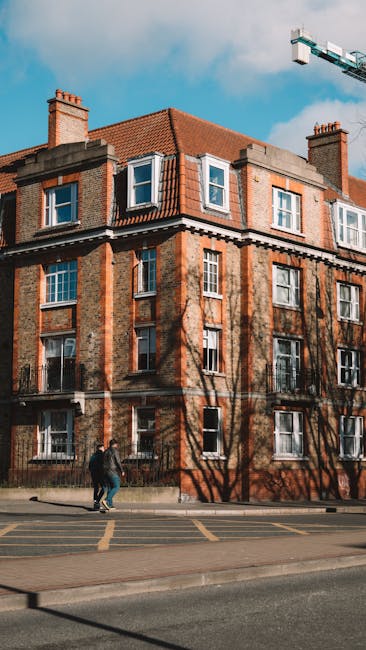 A street scene showing the exterior of several multi-storey residential buildings with brick and concrete facades, balconies, and windows. The road curves slightly and is marked with a large 'SLOW' sign painted in white on the asphalt, with a blue marked parking or loading zone nearby. There is a tall street lamp on the left side of the image, and a few pedestrians are visible walking along the pavement. In the foreground, a man with a moving box wrapped in plastic and cardboard stands on the pavement near the entrance of one building. Man with Van Hampton, a professional removal company, is preparing for a home relocation in Hampton's walk-up buildings, with furniture, boxes, and packing materials visible as part of the loading process inside the property and on the pavement adjacent to a transport van, which is partially out of the frame on the right side. The scene is captured during daylight with overcast skies, utilizing natural lighting to showcase elements of furniture transport, packing, and loading steps involved in house removals.