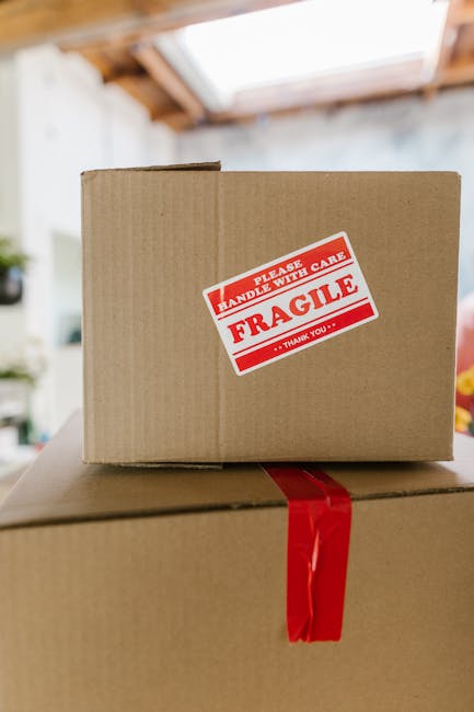 Close-up of two cardboard boxes, one stacked on top of the other, inside a home during a house removal process. The upper box features a red and white 'Fragile' sticker with instructions to handle with care, indicating contents that require delicate handling. The lower box is secured with a red plastic strap around its middle. In the background, part of a room with a skylight window, exposed wooden ceiling beams, and soft natural lighting is visible, suggesting an indoor space prepared for furniture transport. This scene is representative of the packing and loading stages involved in a home relocation, as handled by [COMPANY_NAME], emphasizing careful packing and organizing fragile items for safe transport during a professional removal service.