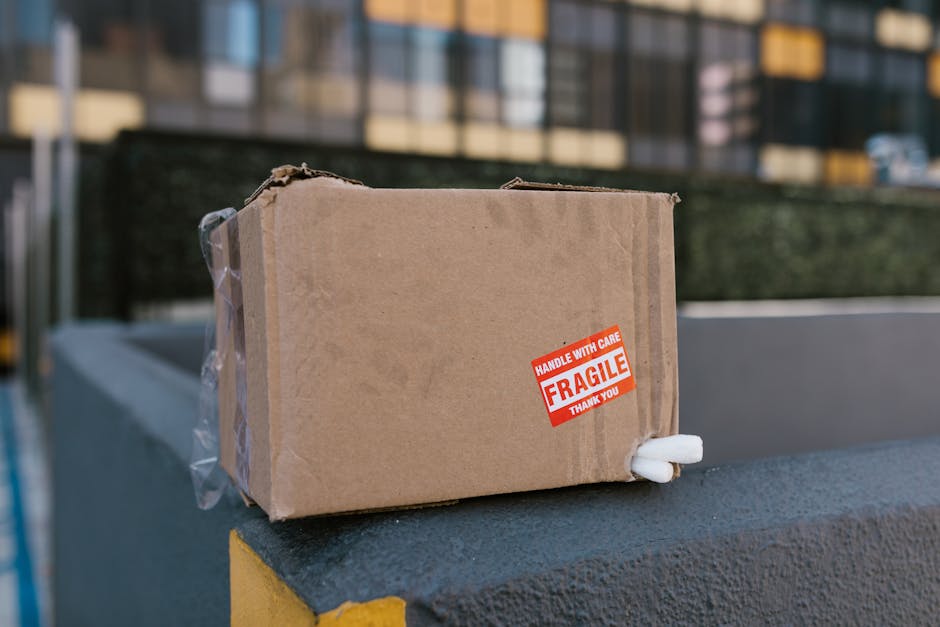 A small cardboard box sealed with clear packing tape is placed on a grey concrete surface outdoors, next to a yellow-painted ledge. The box features a red and white 'FRAGILE' sticker indicating it contains delicate items, and a foam packing piece protrudes from the corner for added protection. In the background, there is an apartment building with large windows and other urban structures, with soft natural lighting illuminating the scene. The setting suggests a home relocation or furniture transport process, possibly being prepared for loading onto a moving van by Man with Van Hampton, specializing in removals. The image highlights careful packing techniques used in house moving, especially for fragile antiques or valuable items, emphasizing the importance of proper packing materials like cardboard boxes, plastic wrap, and foam padding during furniture transport and moving logistics.