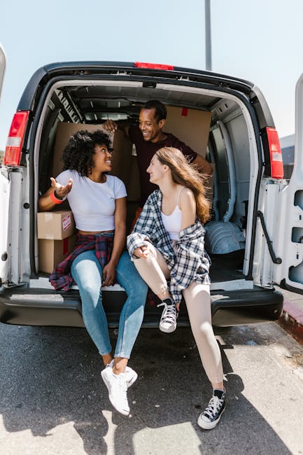 Three young women are sitting inside a moving van parked on the street, engaging in a lively conversation and smiling at each other. The woman on the left, with curly hair, is wearing a white T-shirt,