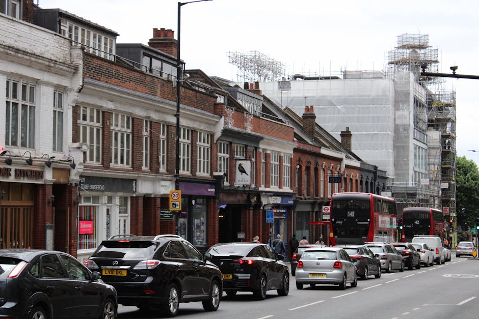 A city street scene during daytime showing a row of traditional brick terraced houses with large multi-pane windows and white decorative trim, situated along a busy pavement. Several cars, including black and silver models, are parked parallel to the kerb, with a black Mercedes-Benz and a grey Fiat among them. Moving inside, a man with a blue shirt is seen carrying a cardboard box wrapped in plastic, while another person nearby is handling a packed cardboard box with packing paper visible. A red double-decker bus is stopped at a bus stop further along the road, with the route number 343 displayed on the front. In the background, a building covered in scaffolding and white protective sheeting indicates ongoing construction or renovation. The street is lit with natural daylight, and the scene captures an active home relocation process involving packing and transport, with the professional services of Man with Van Hampton supporting furniture transport and moving logistics.