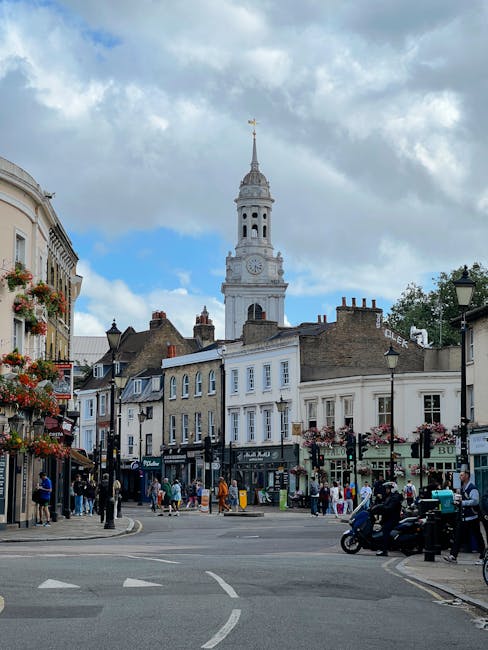 A busy street scene on Hampton Hill High Street featuring a variety of shops and pedestrians walking along the pavement. In the background, a tall, ornate clock tower with a weather vane atop stands prominently against a partly cloudy sky. The street is lined with historic buildings, some with hanging flower baskets and signage for different retail outlets. On the pavement, a few individuals are carrying shopping bags, while others are engaged in conversation or waiting at shop entrances. A group of motor scooters is parked near the curb on the right side, close to a traditional black street lamp. The image illustrates a typical day in a lively, residential shopping area, with ongoing activities related to home and furniture transport, possibly in the midst of house removals or local moving logistics, as exemplified by the context of services like Man with Van Hampton.