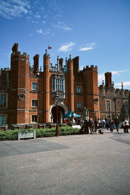 Aerial view of a historic castle surrounded by lush green gardens and trees, with a river flowing nearby and open fields extending into the distance under clear, sunny sky. The castle features multipl