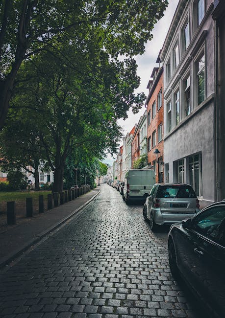 A narrow residential street with brick and stone buildings lining both sides, featuring multiple stories and traditional sash windows. The street is paved with asphalt and has a slight bend, with park