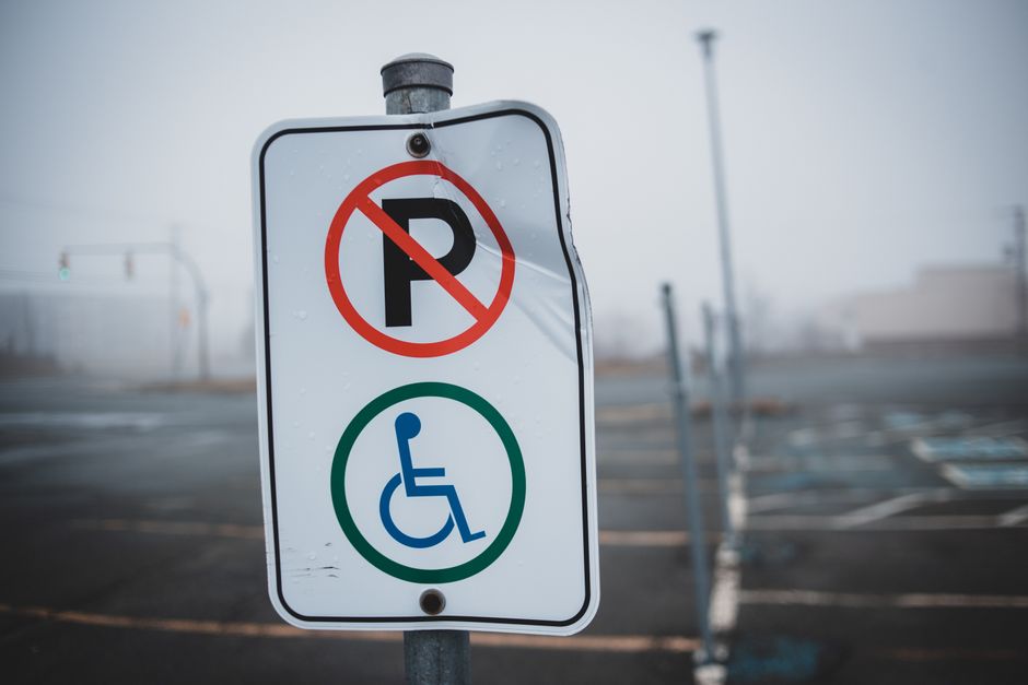 Close-up view of a parking regulation sign positioned on a metal pole in an outdoor parking lot near Hampton Station, HAMPTON. The sign displays a red circle with a diagonal line crossing out the letter 'P', indicating no parking allowed, and a blue wheelchair symbol below, signifying designated accessible parking. The background features an empty parking area with marked parking spaces, some metal barriers, and a cloudy sky, suggesting overcast weather. This image reflects parking restrictions relevant to house removals and furniture transport in the vicinity of Hampton Station, where careful planning of loading zones and accessible parking is essential for efficient home relocation and transportation logistics. The signage supports some aspects of parking rules and can influence moving service planning by informing about designated areas for vehicles involved in removals, as can be organized by companies like Man with Van Hampton.