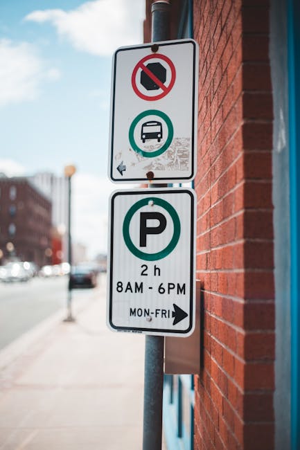 A street photograph showing a metal parking restriction sign affixed to a pole positioned beside a red brick wall. The upper part of the sign features a white background with a red circle and slash over a black no-parking symbol, indicating parking is prohibited in this area. Below that, there is a green circle with a white bus icon, signifying bus restriction. Further down, a large green circle contains a black letter 'P,' with details indicating parking is allowed for a maximum of 2 hours between 8 AM and 6 PM from Monday to Friday. The sign is situated on a sidewalk with a blurred background of parked cars and buildings, suggesting an urban environment. For companies like Man with Van Hampton, this signage indicates parking regulations relevant to local house removals, home relocation, and furniture transport logistics, often requiring awareness of parking rules during loading and unloading processes near Hampton Station.