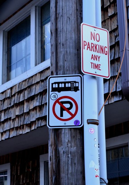 Close-up view of a parking regulation sign positioned on a metal pole in an outdoor parking lot near Hampton Station, HAMPTON. The sign displays a red circle with a diagonal line crossing out the letter 'P', indicating no parking allowed, and a blue wheelchair symbol below, signifying designated accessible parking. The background features an empty parking area with marked parking spaces, some metal barriers, and a cloudy sky, suggesting overcast weather. This image reflects parking restrictions relevant to house removals and furniture transport in the vicinity of Hampton Station, where careful planning of loading zones and accessible parking is essential for efficient home relocation and transportation logistics. The signage supports some aspects of parking rules and can influence moving service planning by informing about designated areas for vehicles involved in removals, as can be organized by companies like Man with Van Hampton.