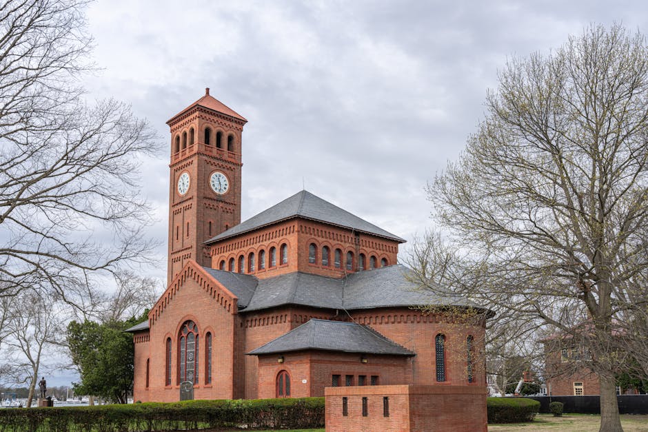 Exterior view of a historic red brick church with a prominent clock tower, situated amidst leafless trees on a cloudy day. The church features arched windows, a sloped slate roof, and decorative brickwork along the roof edges. In the foreground, a paved area and a small brick boundary wall are visible. The scene depicts a peaceful setting typical of a town centre or residential area, with the church’s architecture highlighting its traditional design. Man with Van Hampton's removal team may conduct home relocation or furniture transport activities nearby, represented by loading and unloading operations involving packing materials and moving equipment. The image emphasizes the careful handling of furniture and boxes during packing and loading, set against the backdrop of a well-maintained property exterior, which is relevant to house removals and moving logistics services.