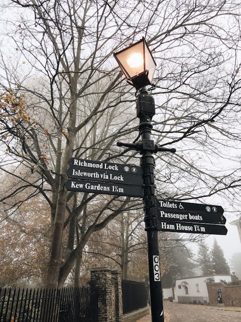 A black lamppost with a lit lantern on top, situated on a pavement next to a leafless tree. Attached to the lamppost are multiple directional signs indicating nearby locations, including 'Richmond Loc