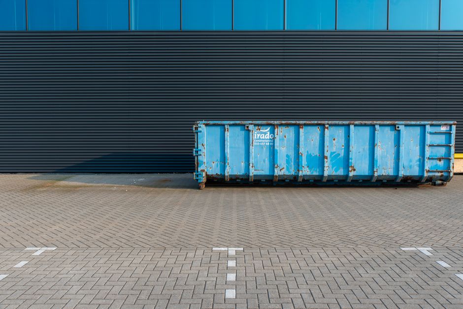 A row of four large white plastic waste bins with closed lids, positioned on a paved surface outside of a property. To the left, there is a dark grey recycling bin labeled 'PAPER' and a similar bin la