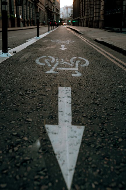 A close-up view of a designated bike lane on a city street, featuring a white bicycle symbol painted on the asphalt with a large downward-pointing arrow directly below it. The lane is separated from the vehicle lanes by short black posts with reflective markings, and the pavement surface shows small gravel and textured concrete markings. In the background, there are tall, historic buildings lining the street, and traffic lights are visible at the intersection ahead. The scene is lit with natural daylight, capturing the urban environment that a home relocation or furniture transport company like Man with Van Hampton might encounter when navigating narrow streets during moving and packing activities associated with house removals.