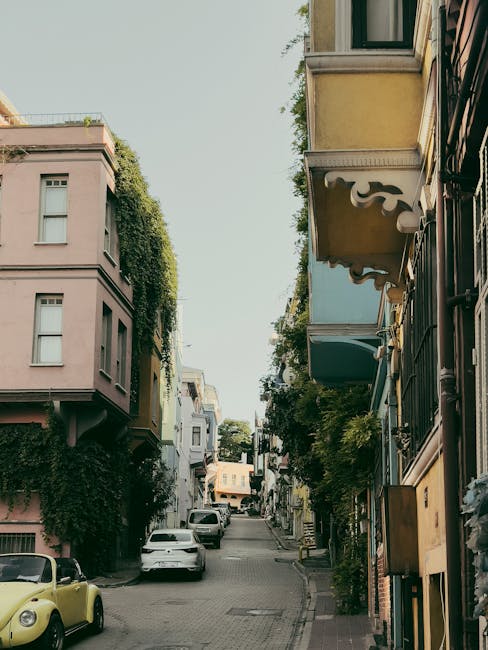 The image shows a narrow, cobblestone street in a residential area with multi-storey buildings on either side, featuring various architectural styles and pastel-colored facades. Some building exteriors are decorated with greenery, such as ivy climbing up the walls. The street is lined with parked cars, including a distinctive yellow vintage car on the left side, and a white vehicle further along the street. The scene is outdoors during daylight hours, with natural light illuminating the area and casting soft shadows. There are no visible people in the image. This setting is typical for house removals and furniture transport in confined urban streets, and the image reflects the type of environment where careful planning for parking and space management is essential for home relocation services offered by Man with Van Hampton.